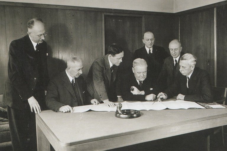 Schwarz-weiß Archiv-Aufnahme von sieben älteren Männern in schwarzen Anzügen bei der Unterzeichnung von Verträgen auf einem Tisch in einem Raum mit Holzverkleidung