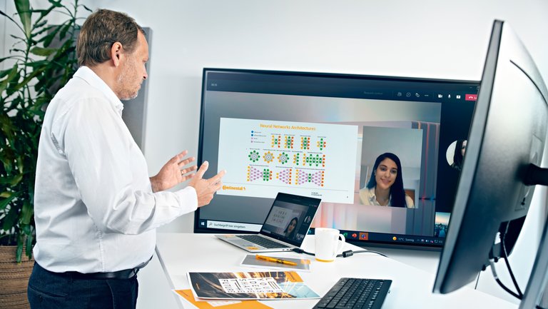 An older man in a white shirt stands at a standing workstation with keyboard, screen and laptop and looks at a TV screen on which a video conference is taking place
