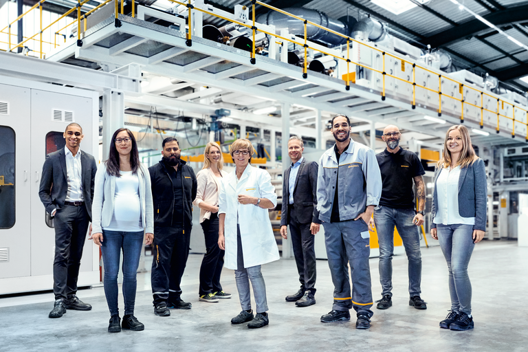 Zwei Männer in Anzügen, zwei Männer in Arbeitskleidung, eine Frau im Kostüm, eine Frau im Kittel, eine Frau und ein Mann in legerer Arbeitskleidung und eine schwangere Frau stehen in einer Produktionshalle und lächeln