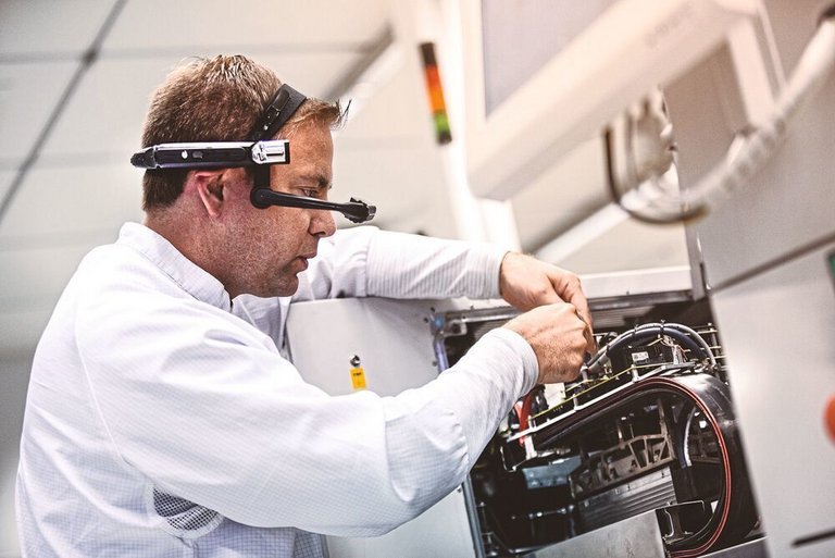 A man in a white shirt and a magnifying glass on his head plugs cables together on a machine