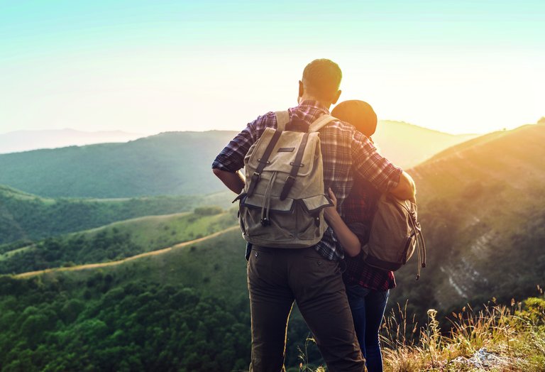 A man and a woman in hiking outfits and hiking backpacks embrace and look into a valley during sunset.