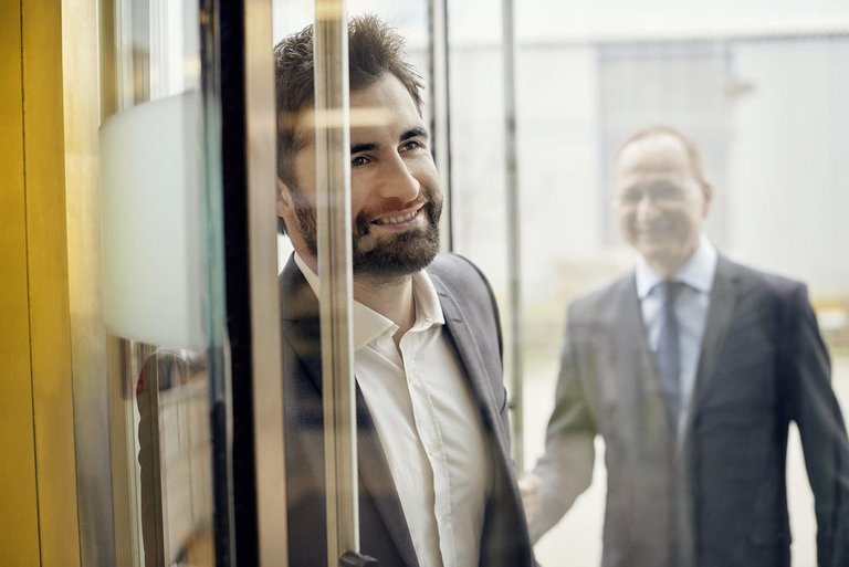 Doi bărbați în costume intră într-o sală pe o uşă de sticlă