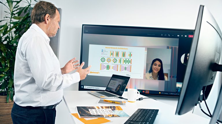 Ein älterer Mann in weißem Hemd steht an einem Steharbeitsplatz mit Tastatur, Bildschirm und Laptop und schaut auf einen TV-Screen, auf dem eine Videokonferenz stattfindet