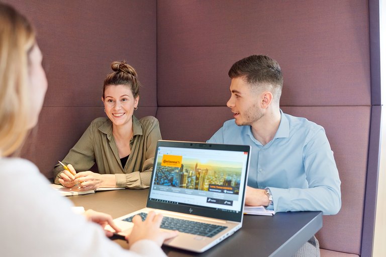 Zwei junge Frauen und ein junger Mann sitzen an einem Tisch mit Laptop und lächeln