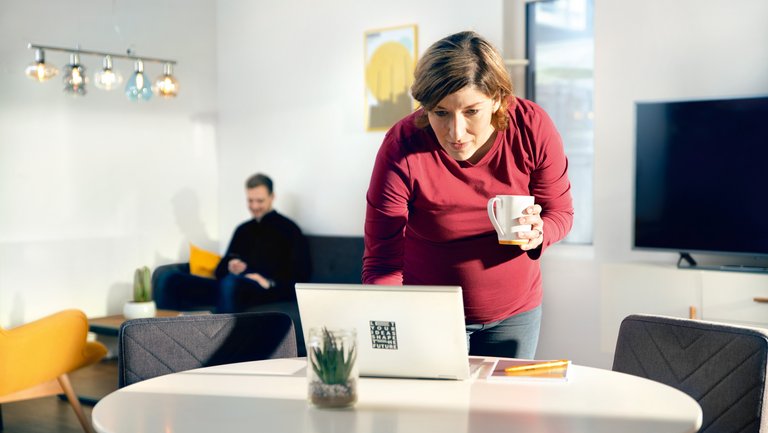 A pregnant woman in a red jumper holds a coffee mug and types something on a laptop while a man sits on a sofa in the background