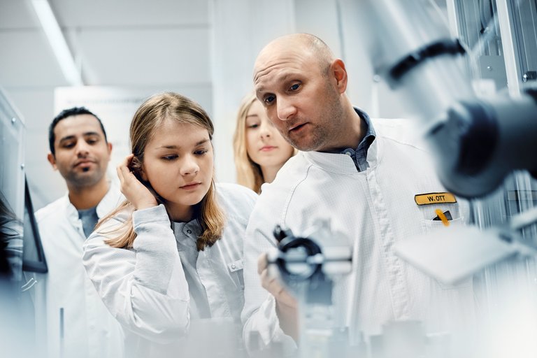 A bald man shows two young women and a young man a device in the laboratory 