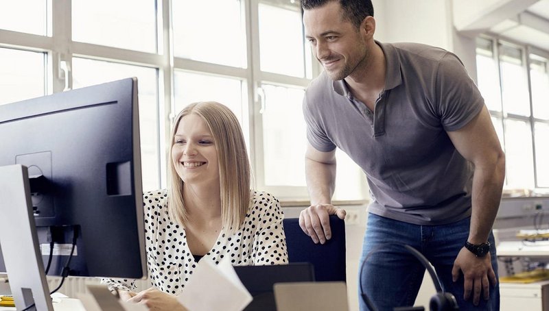 A young woman sits in front of a computer screen talking with the man standing behind her