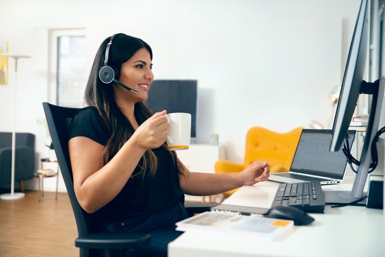 A young woman with dark hair in a black T-shirt wears a headset and holds a coffee pot while sitting at her home office desk.