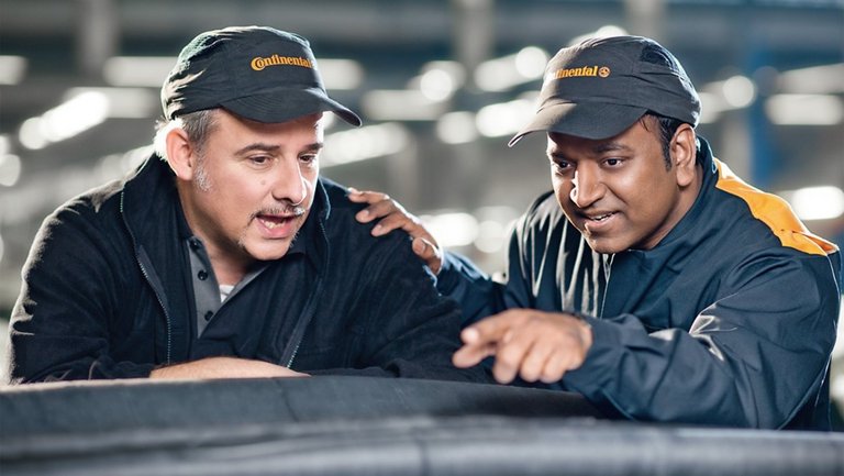 Ein älterer und ein jüngerer Mann in Arbeitskleidung und Caps mit Continental-Logo schauen auf ein Förderband