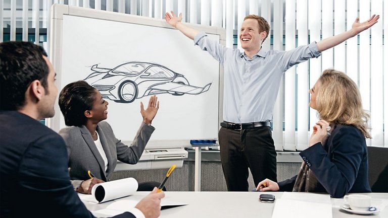 Ein Mann steht vor einem Whiteboard, auf dem ein Auto mit Flügeln abgebildet ist und streckt beide Arme glücklich von sich, während zwei Frauen und ein Mann ihn anschauen
