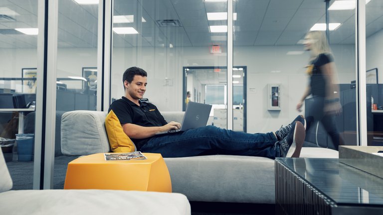 Ein Mann in einem schwarzen Continental-Poloshirt sitzt mit hochgelegten Beinen auf einem Sofa in einem Glaskasten und tippt auf dem Laptop auf seinem Schoß