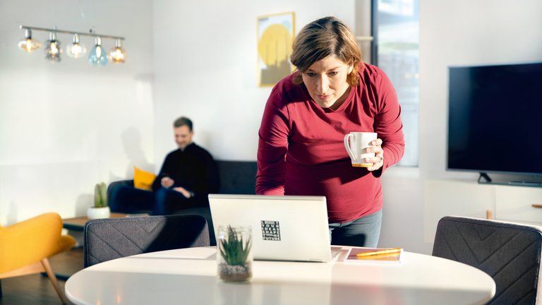 Eine schwangere Frau in rotem Pullover hält einen Kaffeebecher und tippt etwas am Laptop, während ein Mann im Hintergrund auf einem Sofa sitzt