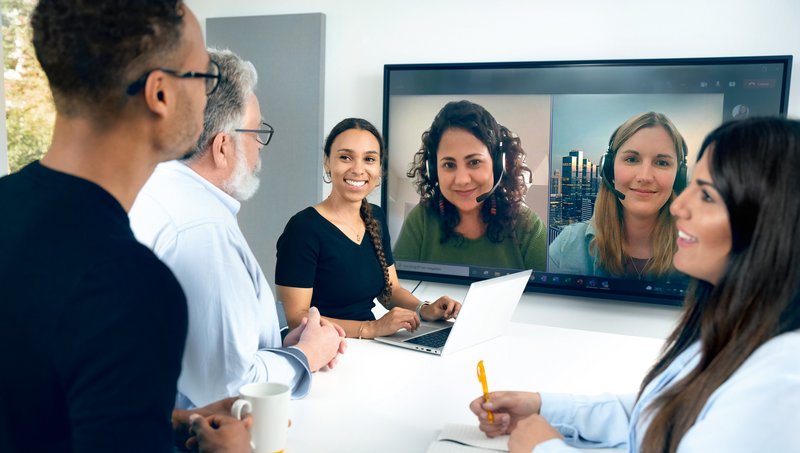 Zwei junge Frauen und zwei Männer stehen an einem Stehtisch und blicken auf einen Fernseher, auf dem zwei Frauen mit Headset in einer Videokonferenz abgebildet sind