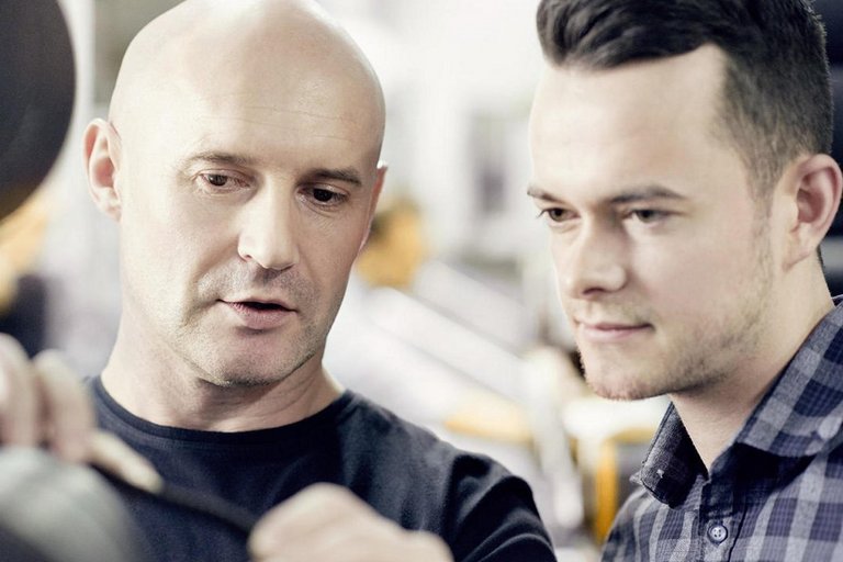 Ein Mann mit Glatze erklärt einem jüngeren Mann in Karohemd ein technisches Produkt