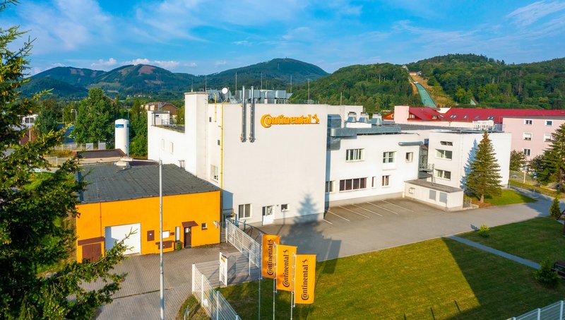 Pohled z dronu na bíložluté budovy areálu Continental ve Frenštátu pod Radhoštěm se zelenými kopci v pozadí