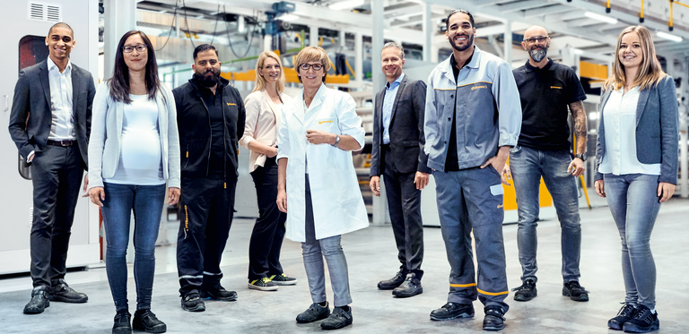 Two men in suits, two men in work clothes, a woman in a suit, a woman in a lab coat, a woman and a man in casual work clothes, and a pregnant woman standing in a production facility smiling