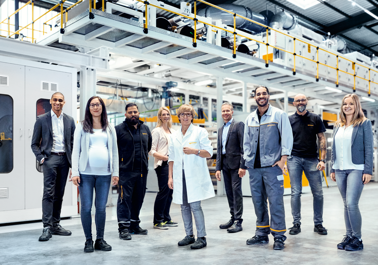 Two men in suits, two men in work clothes, a woman in a suit, a woman in a smock, a woman and a man in casual work clothes and a pregnant woman stand in a production hall and smile