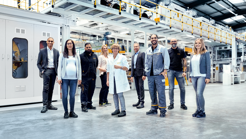 Two men in suits, two men in work clothes, a woman in a suit, a woman in a lab coat, a woman and a man in casual work clothes, and a pregnant woman standing in a production facility smiling 
