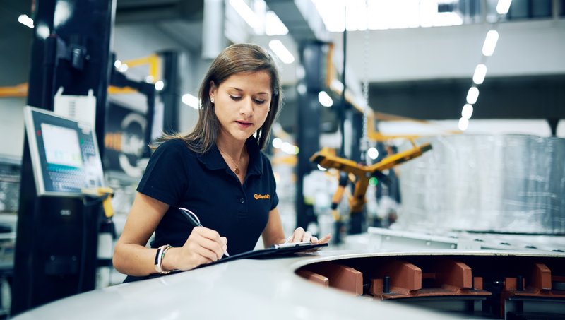 A young woman in a polo shirt stands in a factory and notes down something