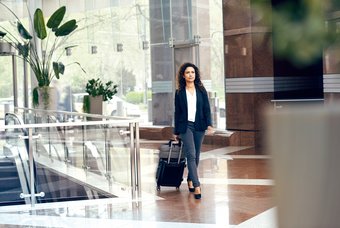 A woman in a jacket, jeans and pumps pulls a trolli behind her as she walks through a reception hall with an escalator
