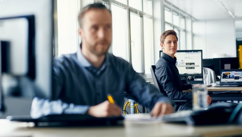 Two young men are sitting at their desks in front of their computer in an open office