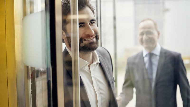 Doi bărbați în costume intră într-o sală pe o uşă de sticlă