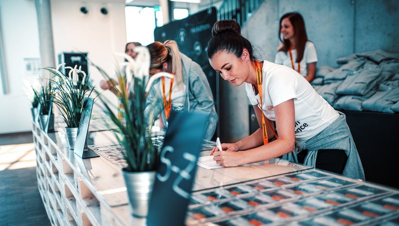 Eine junge Frau in weißem T-Shirt und Schlüsselband um den Hals steht an einem Messestand und notiert etwas auf einem Zettel