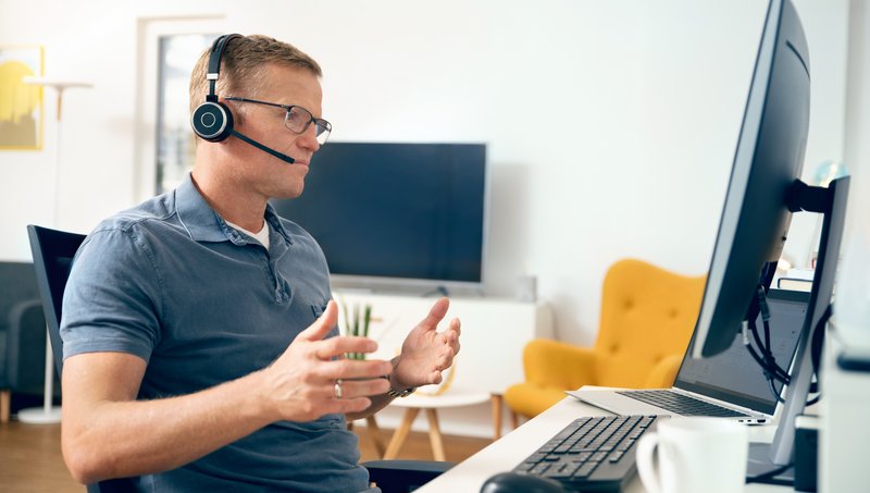 Ein Mann mit Brille, Polohemd und Headset sitzt gestikulierend vor einem Arbeitsplatz