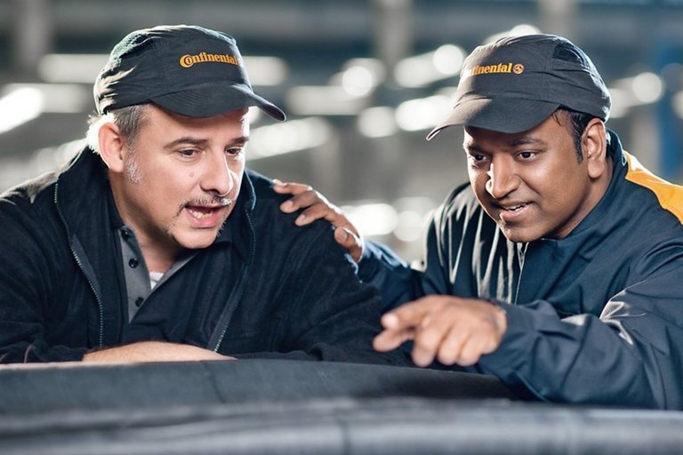 An older and a younger man in work clothes and caps with Continental logo look at a conveyor belt
