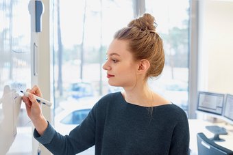 eine junge Frau mit roten Haaren und rotem Lippenstift steht vor einem White Board und schreibt etwas daran