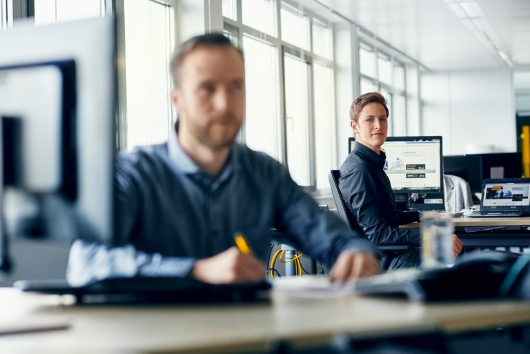 A man at a desk in the background looks at a man at a desk in the foreground