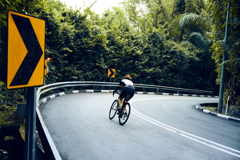 A racing cyclist turns into the bend of a dual carriageway bordered by a crash barrier, road signs trees and bushes