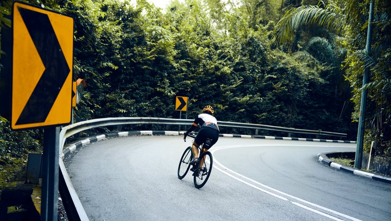 Ein Rennradfahrer biegt in die Kurve einer Schnellstraße ein, die von einer Leitplanke, Straßenschildern Bäumen und Büschen eingegrenzt wird