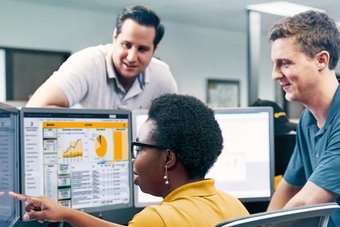 Eine mittelalte Frau sitzt vor drei PC-Bildschirmen, die Statistiken abbilden, und zeigt zwei Männern auf dem linken Display etwas