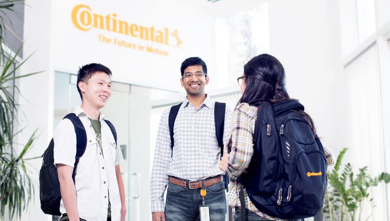 Zwei junge Männer und eine junge Frau und legerer Businesskleidung stehen in einer Empfangshalle vor einem Fahrstuhl und unterhalten sich