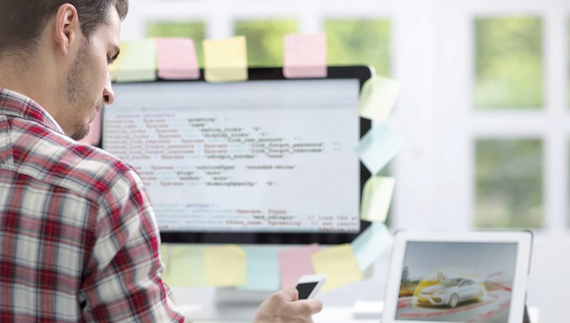 Junger Mann programmiert eine App am Computer und überprüft das Ergebnis am Smartphone.