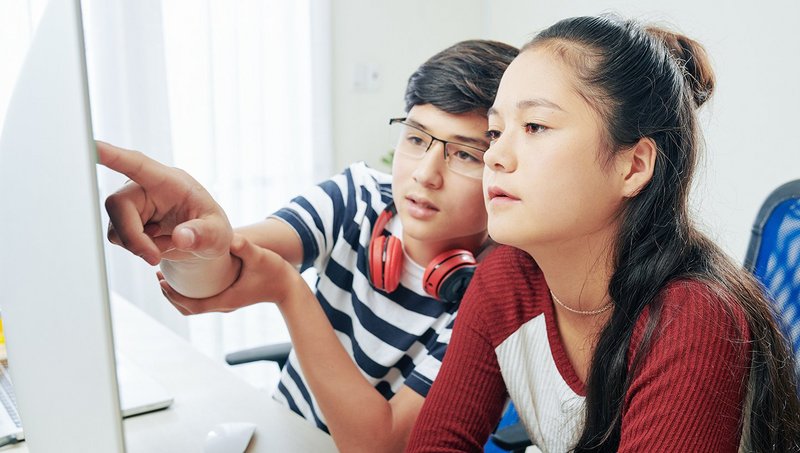 Ein Junge mit Kopfhörern und gestreiftem T-Shirt und ein Mädchen in rotem Pullover schauen auf einen Bildschirm