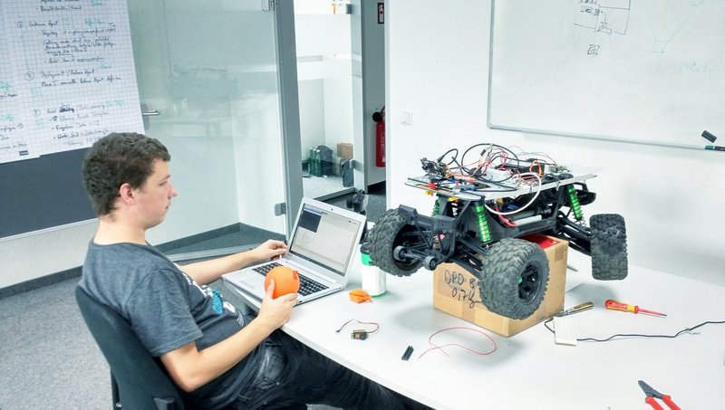 A young man sits at a desk in front of his laptop, next to him an RC car