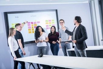Three young women and two young men stand in front of a white board and look towards a middle-aged man in a jacket who explains the contents of the white board