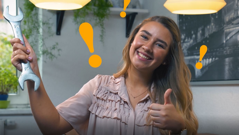 Eine junge Frau hält den Daumen hoch und einen Maulschlüssel in der anderen Hand, während sie lächelt und orangene Ausrufezeichen um sie herumfliegen