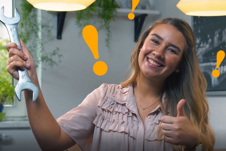 Eine lächelnde junge Frau, die einen Maulschlüssel hält und den Daumen in die Kamera hält, während orangene Ausrufezeichen um ihren Kopf schweben in einem Büro-Setting