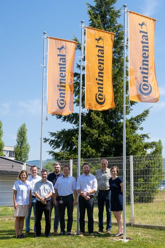 Osm členů HR týmu Continental Frenštát pod Radhoštěm v prosluněném dni na trávníku pod oranžovými vlajkami Continentalu
