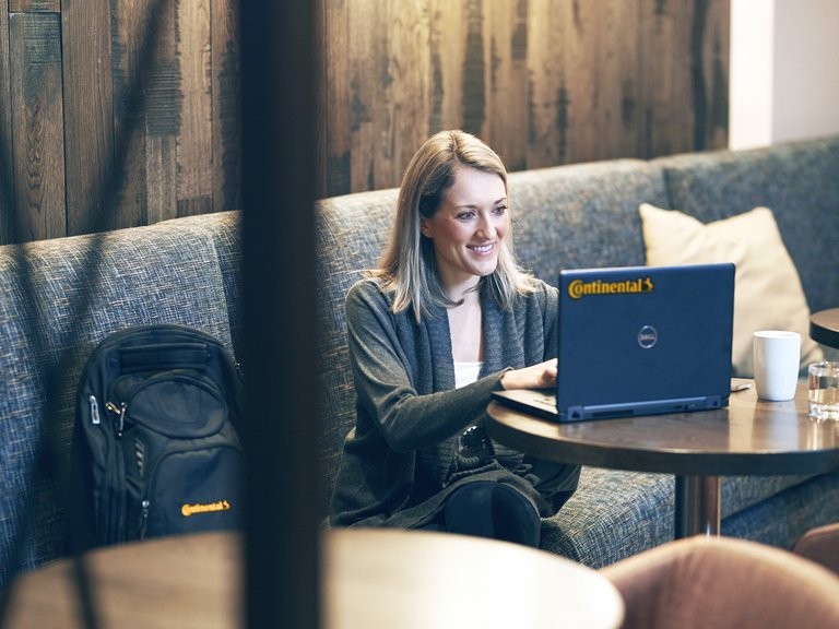 Eine Frau sitzt auf einem Sofa, neben ihr ein Rucksack mit Continental-Logo, und arbeitet lächelnd an einem Laptop mit Continental-Logo