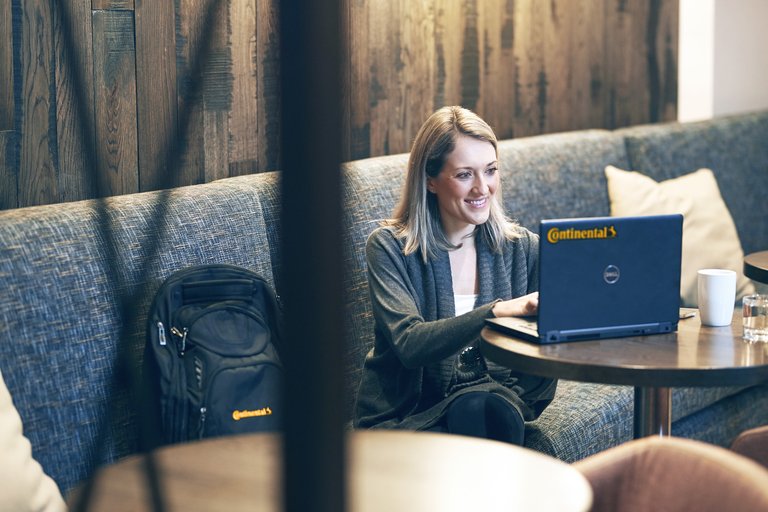 A woman sits on a sofa, next to her a backpack with Continental logo, and works smiling on a laptop with Continental logo