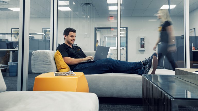 A man in a black Continental polo shirt sits with his legs up on a sofa in a glass box and types on the laptop on his lap